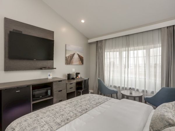 A modern hotel room featuring a bed, flat-screen TV, artwork, curtains over a large window, a desk, two chairs, and a small round table.