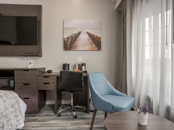 This image shows a modern hotel room with a TV, desk, chair, and decor. A window with curtains is on the right, letting in natural light.