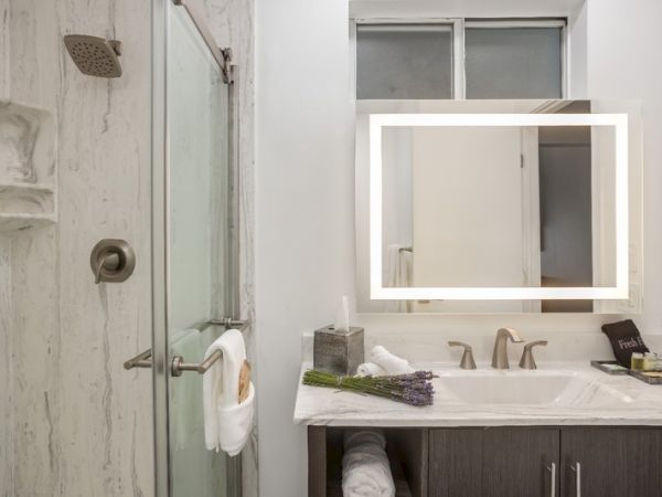 A modern bathroom with a shower, illuminated mirror, sink, countertop items, and fresh towels.