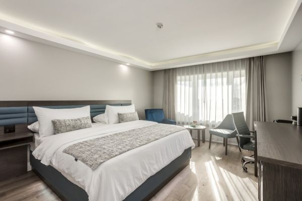 The image shows a modern hotel room with a large bed, a desk, a chair, a window with curtains, and minimalistic decor.