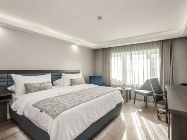 The image shows a modern hotel room with a large bed, a desk, a chair, a window with curtains, and minimalistic decor.