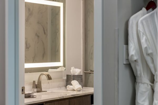 A bathroom with a lit mirror, sink with a faucet, rolled towels, and tissue box is visible. Two white bathrobes hang on a hook inside the doorway.