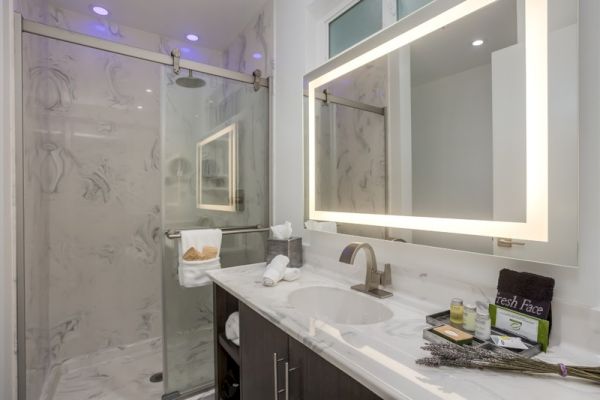 The image shows a modern bathroom with a marble shower, illuminated mirror, and a sink with toiletries and towels neatly arranged on the countertop.