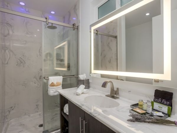 The image shows a modern bathroom with a marble shower, illuminated mirror, and a sink with toiletries and towels neatly arranged on the countertop.