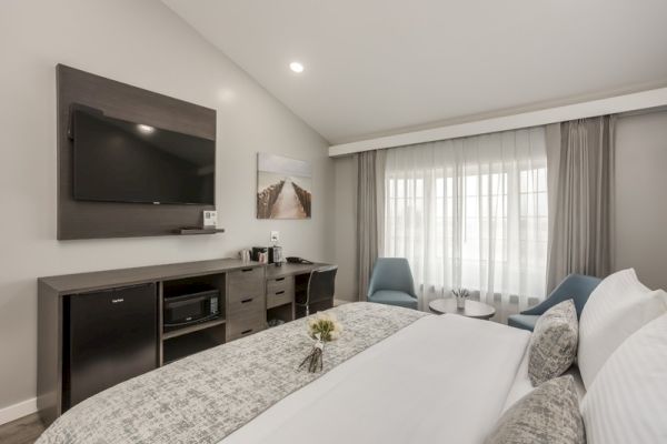 The image shows a modern hotel room with a bed, TV, and a small sitting area with two chairs and a table near a large window with curtains.