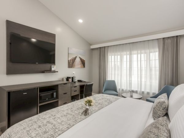The image shows a modern hotel room with a bed, TV, and a small sitting area with two chairs and a table near a large window with curtains.