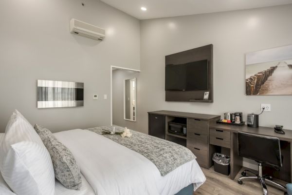 A modern hotel room with a bed, TV, desk, coffee maker, and wall art. The room has gray walls and a sleek, minimalist style.