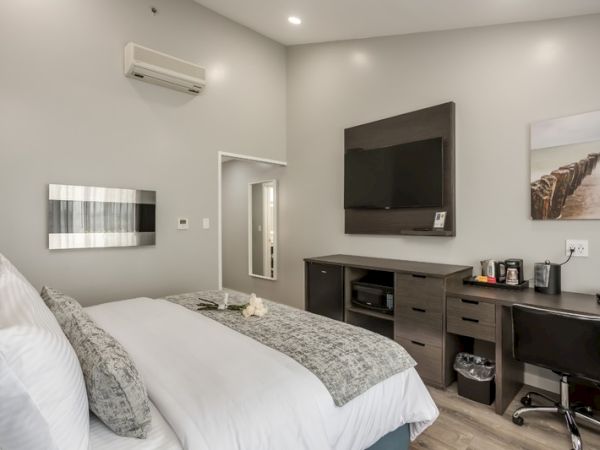 A modern hotel room with a bed, TV, desk, coffee maker, and wall art. The room has gray walls and a sleek, minimalist style.