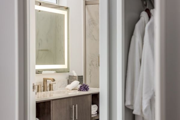 A modern bathroom features a large illuminated mirror, marble countertop, gold faucet, decorative items, and white robes hanging on a rack.