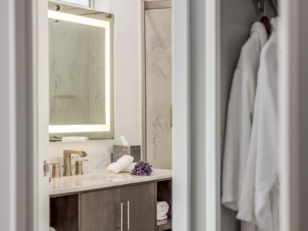 A modern bathroom features a large illuminated mirror, marble countertop, gold faucet, decorative items, and white robes hanging on a rack.
