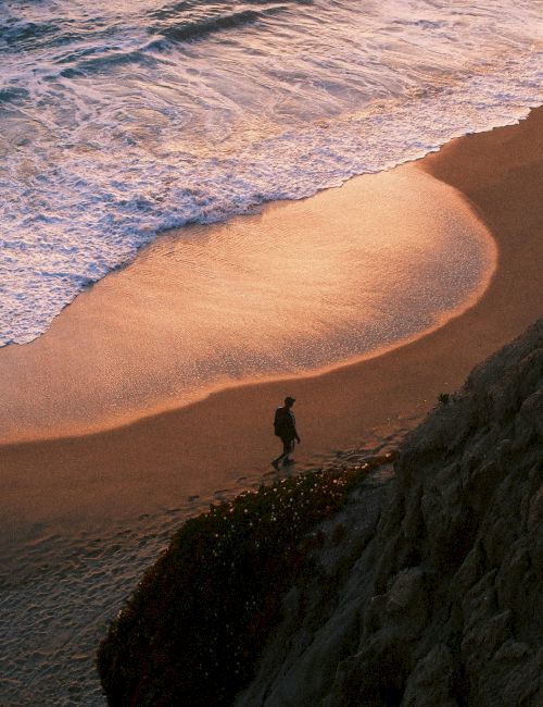 A person is walking along a sandy beach near the shoreline during sunset, with waves gently crashing against the shore.