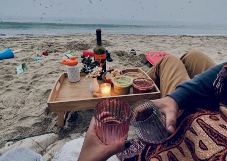 A cozy beach picnic with a tray holding food, flowers, and a wine bottle. Two people toasting with drinks, wrapped in blankets by the shore.