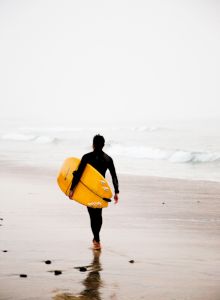 A person in a wetsuit walking along the beach towards the ocean, holding a yellow surfboard. The weather appears foggy, and the waves are calm.