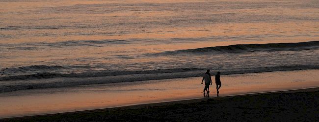 A tranquil sunset view over the ocean with two people walking on the beach in the foreground, and an island visible in the distance.