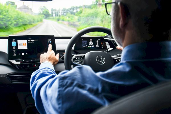 A person is driving a car, interacting with the touchscreen infotainment system. The view is from the backseat, focusing on the driver and dashboard.