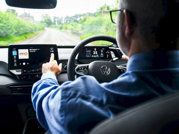 A person is driving a car, interacting with the touchscreen infotainment system. The view is from the backseat, focusing on the driver and dashboard.