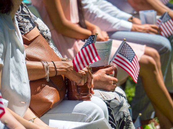 People seated, holding small American flags, possibly during a patriotic event or ceremony, with sunlight highlighting the scene.