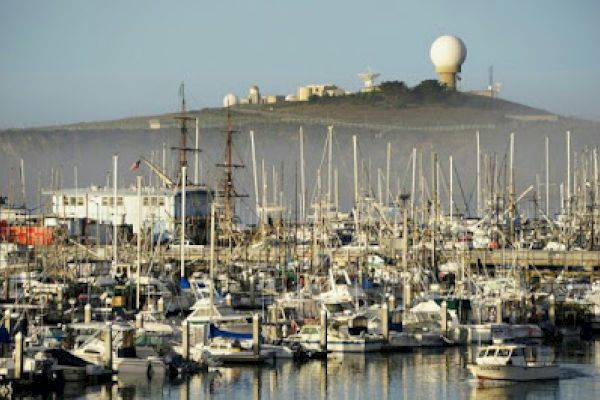 This image shows a marina filled with numerous boats, with a hill in the background featuring a large radar or observation dome structure.