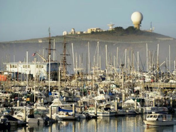 This image shows a marina filled with numerous boats, with a hill in the background featuring a large radar or observation dome structure.