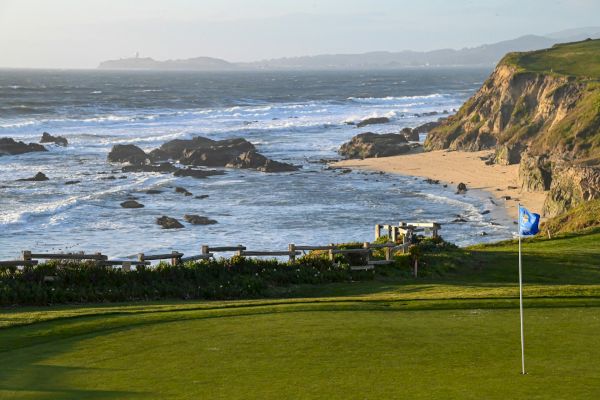 This image shows a golf course with a flag in the foreground, a rocky coastline, and waves crashing on the shore under a partly cloudy sky.