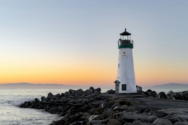 A lighthouse stands on a rocky pier by the ocean at sunrise or sunset, with calm waters and distant mountains visible on the horizon.