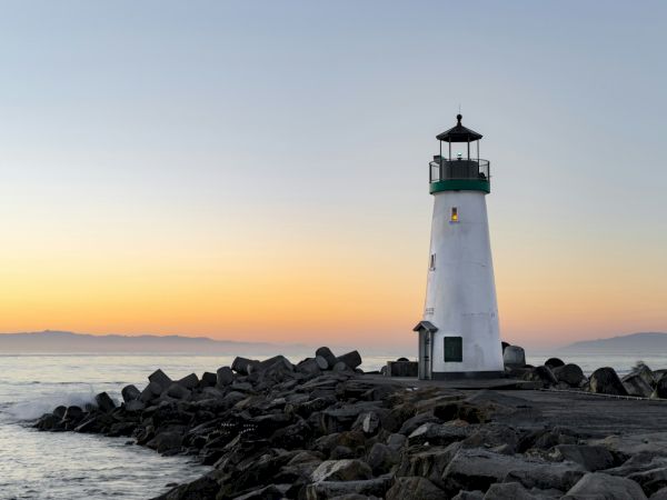 A lighthouse stands on a rocky pier by the ocean at sunrise or sunset, with calm waters and distant mountains visible on the horizon.