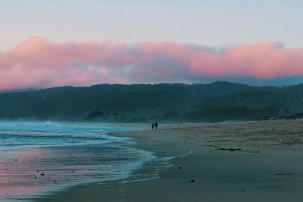 A serene beach scene at sunrise or sunset with a pastel pink sky, waves crashing on the shore, and two people walking along the sandy beach.