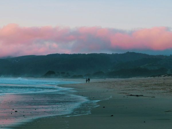 A serene beach scene at sunrise or sunset with a pastel pink sky, waves crashing on the shore, and two people walking along the sandy beach.