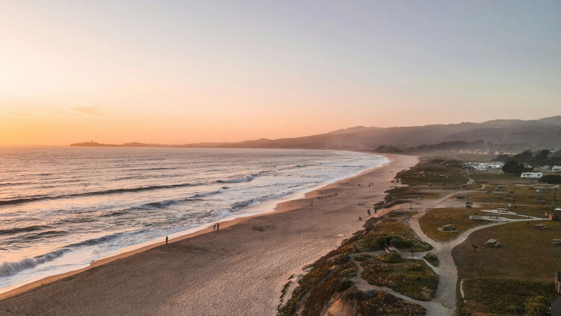 A serene beach at sunset with gentle waves, a sandy shore, and distant hills. People stroll along the beach and walking paths on a clear evening.