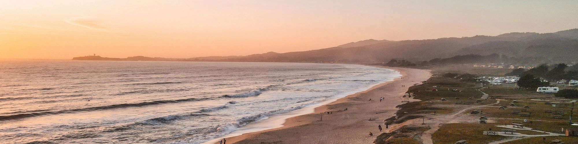 A serene beach at sunset with gentle waves, a sandy shore, and distant hills. People stroll along the beach and walking paths on a clear evening.