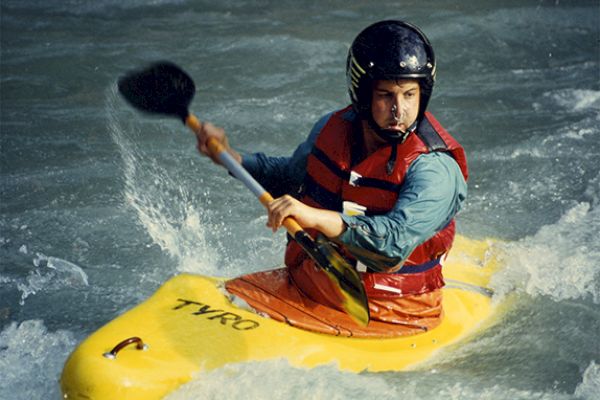 A person is kayaking in turbulent waters, wearing a helmet and life vest, while paddling a yellow kayak.