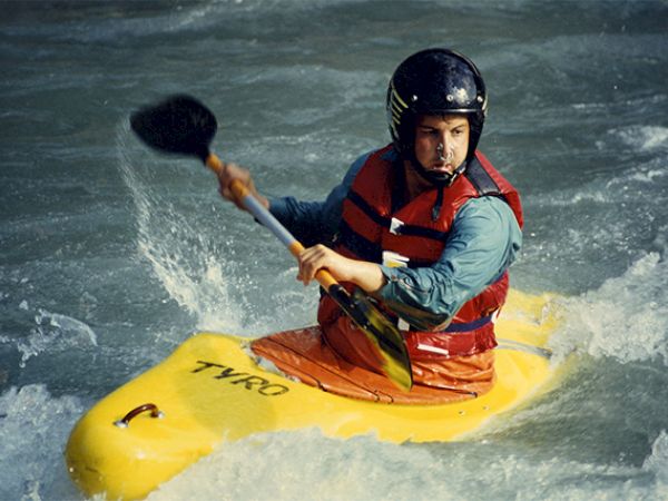 A person is kayaking in turbulent waters, wearing a helmet and life vest, while paddling a yellow kayak.
