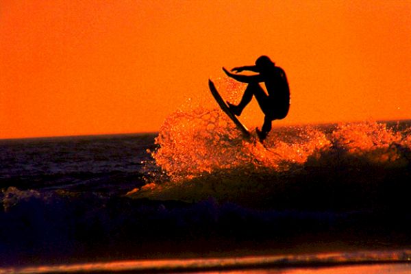 A person is surfing on a wave against a stunning orange sunset backdrop, with water spraying around them as they catch air.