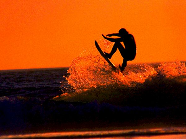 A person is surfing on a wave against a stunning orange sunset backdrop, with water spraying around them as they catch air.