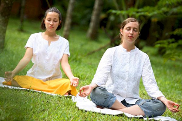 Two people are sitting on the grass in a tranquil environment, meditating with their eyes closed, wearing casual white tops.