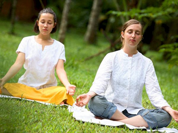 Two people are sitting on the grass in a tranquil environment, meditating with their eyes closed, wearing casual white tops.