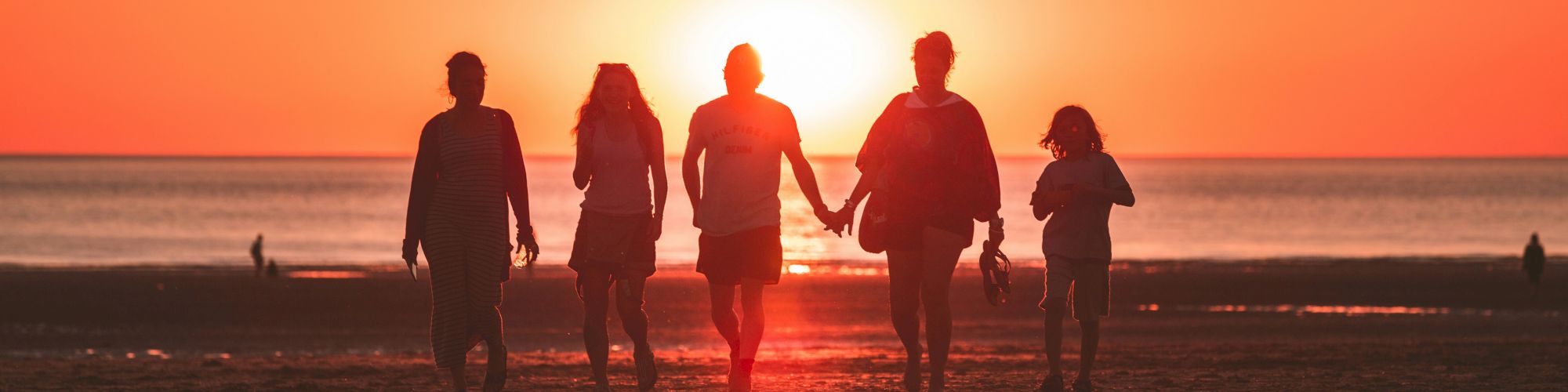 A group of five people walks hand in hand on a beach during sunset, with the bright sun setting over the horizon.