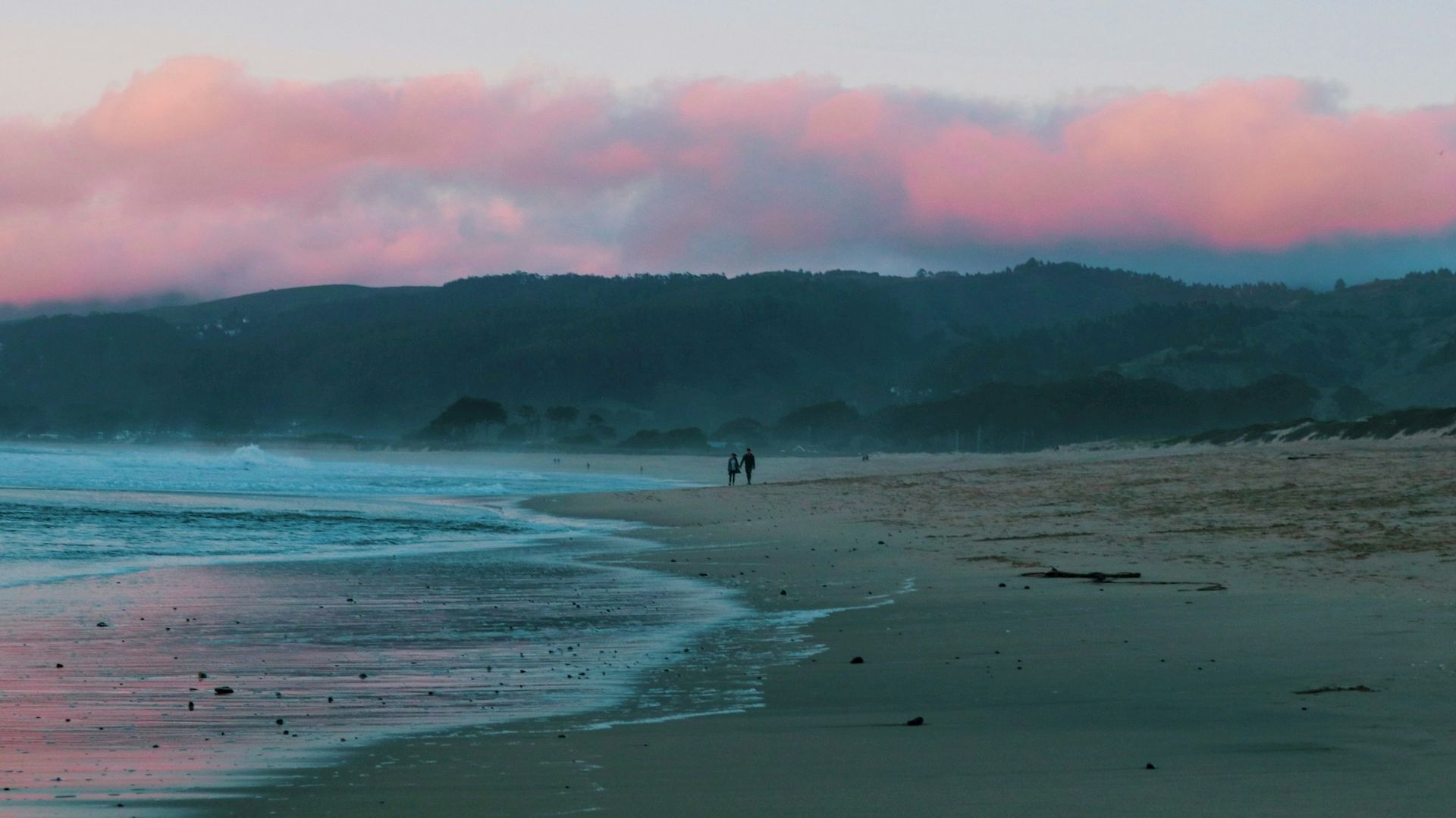 The image shows a serene beach at sunset with two people walking along the shore and a backdrop of pink clouds over distant hills.
