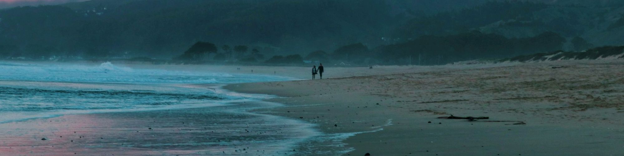 The image shows a serene beach at sunset with two people walking along the shore and a backdrop of pink clouds over distant hills.