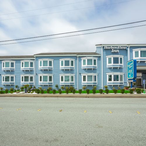 A two-story blue building labeled as a Harbor View Inn along a sidewalk with a parking lot and some landscaped plants in front of it.