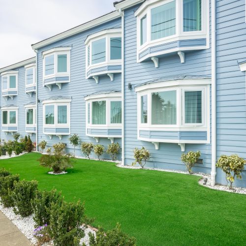 A light blue building with multiple white-framed windows, neat green grass, and bushes along a paved walkway.