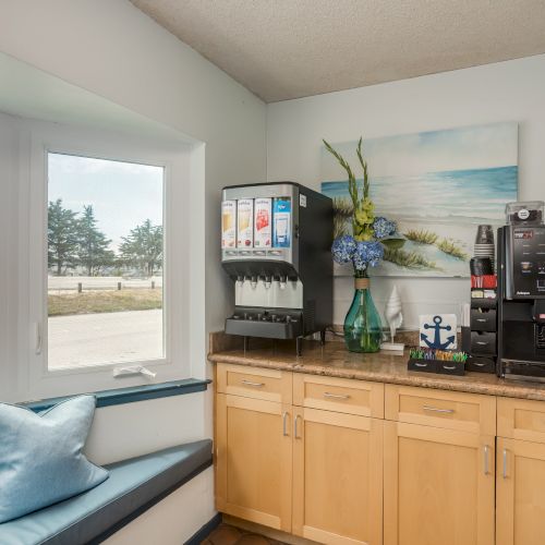 A corner with a drink dispenser, coffee machine, decorative vase, and a coastal-themed painting; next to a window seat with a blue pillow.