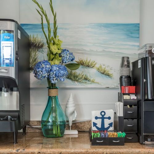 The image shows a drink station with a soda dispenser, a coffee machine, cups, and supplies, set against a nautical-themed background with a beach painting.