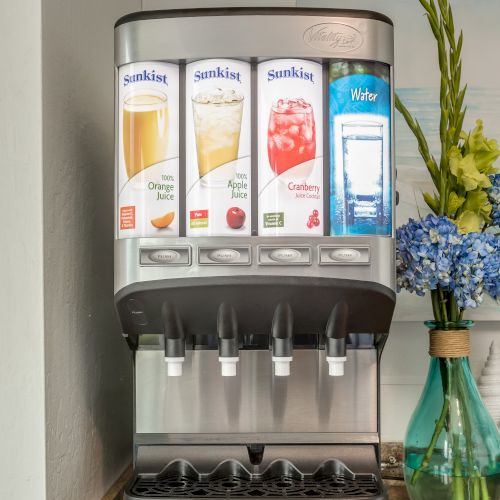 The image shows a beverage dispenser with drink options like orange juice, apple juice, cranberry juice, and water, next to a floral arrangement.