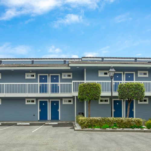 The image shows a two-story motel with blue doors, outdoor staircases, a parking lot, and manicured greenery under a clear blue sky.