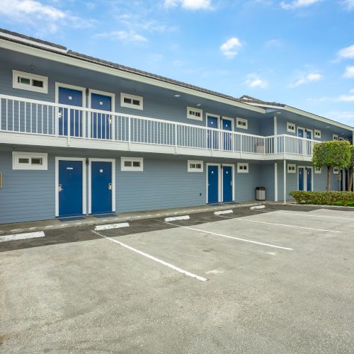 The image shows a two-story motel with blue doors, a gray exterior, and an empty parking lot on a clear day, ending the sentence.