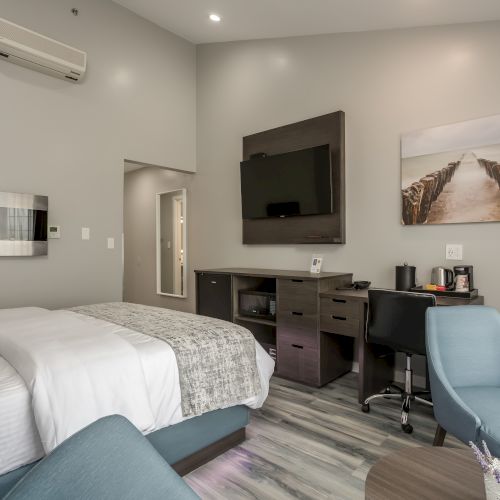 A modern bedroom with a bed, wall-mounted TV, desk, chairs, artwork, air conditioning unit, and kitchenette area. Cozy and neatly arranged.