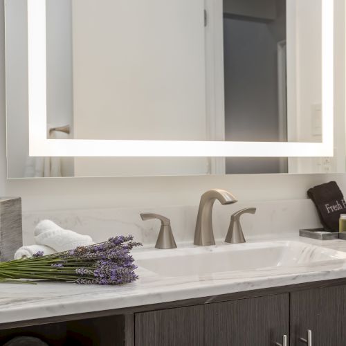 A bathroom sink with a lit mirror above it, a lavender bouquet, tissue box, towel, and a soap dish and container are on the counter.