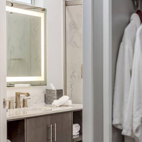 The image shows a modern bathroom with a backlit mirror, a sink with gold faucets, fluffy robes hanging, and a marble shower in the background.