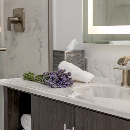 Modern bathroom with marble surfaces, a sink with a brass faucet, a mirror with lighting, a glass shower door, and lavender flowers on the counter.